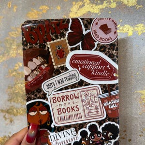 May include: A Kindle case with a leopard print design, adorned with stickers. Stickers include text that says "BRATZ", "BORROW MORE BOOKS", "DIVINE FEMININE", and "READING FUEL". A hand with a red and orange gradient nail polish is holding the case.