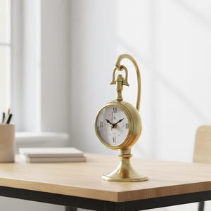 May include: A gold-colored, vintage-style desk clock with a round face and Roman numeral hour markers. The clock has a curved handle and sits on a pedestal base. The clock is on a light-colored wooden desk.