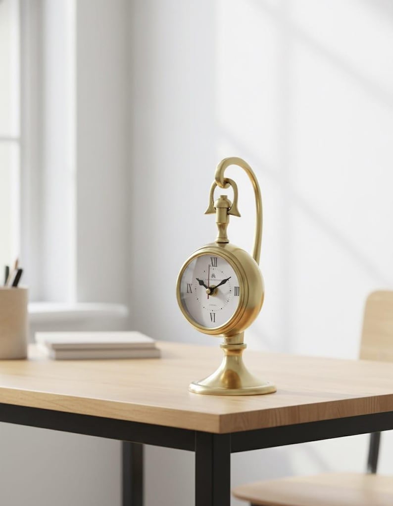 May include: A gold-colored, vintage-style desk clock with a round face and Roman numeral hour markers. The clock has a curved handle and sits on a pedestal base. The clock is on a light-colored wooden desk.