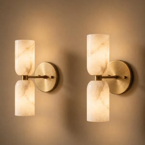 May include: Two wall sconces with a contemporary design. Each sconce has a cylindrical, marble-effect shade in cream, connected to a brass-coloured circular base. The sconces are mounted on a neutral-toned wall, creating a warm, inviting ambience.