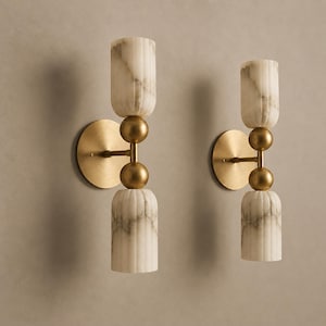 Op de afbeelding: Een paar wandlampen met ronde messing achterplaten en horizontale armen verbonden met verticale structuren. Elk armatuur bevat twee gecanneleerde cilindrische kappen van wit marmer, gescheiden door twee gestapelde messing bollen in het midden.