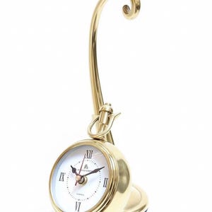 May include: A gold-colored desk clock with a round face and Roman numeral hour markers. The clock has a curved, ornate stand and a small base. The clock face is white with black hands and the brand name "London" is visible.
