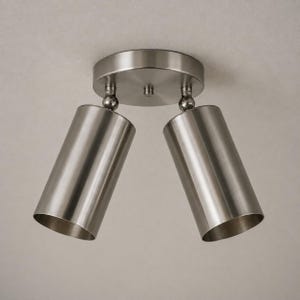Puede incluir: Un foco de techo de níquel cepillado con dos cabezales de luz cilíndricos ajustables. Los cabezales de luz están angulados hacia abajo y conectados a una base circular. El accesorio está diseñado para iluminación direccional.