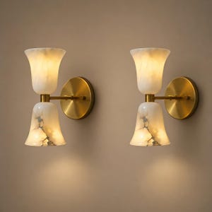 Pode incluir: Duas arandelas de parede com um design de sino duplo. As arandelas apresentam uma sombra de vidro branco marmorizado e uma base de metal cor de latão. São montadas em uma parede de cor neutra, criando um ambiente acolhedor.