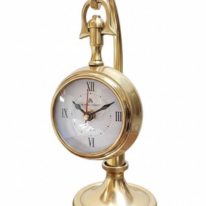 May include: A gold-colored desk clock with a round face and Roman numerals. The clock has a curved handle and a circular base. The clock face is white with black hands and the text "49 Bond Street".
