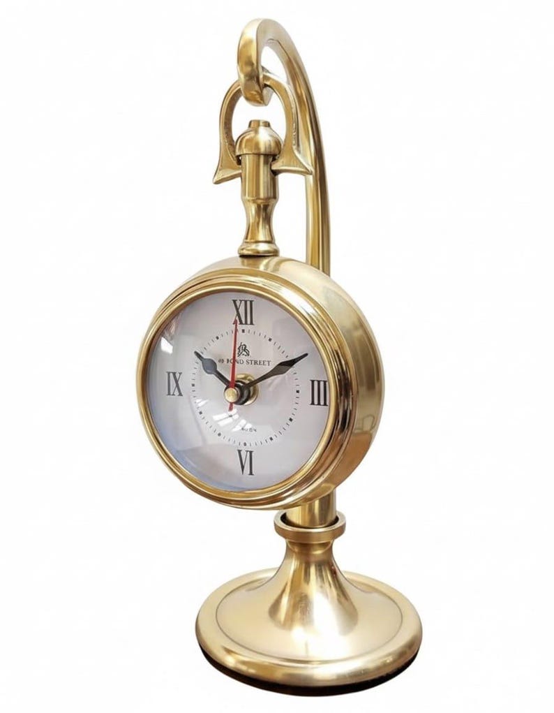 May include: A gold-colored desk clock with a round face and Roman numerals. The clock has a curved handle and a circular base. The clock face is white with black hands and the text "49 Bond Street".