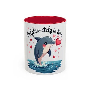 May include: White ceramic mug with a red interior featuring a cartoon dolphin jumping out of water. The dolphin is gray with a pink blush, surrounded by hearts and the text "Dolphin-ately in love."