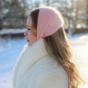 May include: A woman wearing a pale pink knitted head covering and a white faux fur coat. The woman has long, wavy blonde hair. The background is a snowy landscape with trees and sunlight.