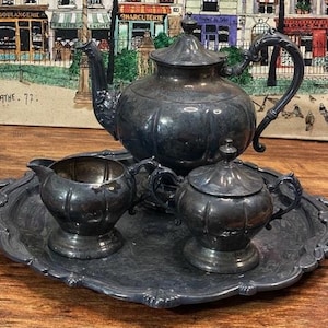 May include: A vintage silver tea set, including a teapot, creamer, and sugar bowl, sits on a decorative tray. The set has a dark, tarnished finish and ornate handles. The background features a framed print of a European street scene.