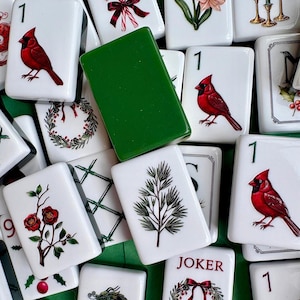 May include: A collection of rectangular mahjong tiles featuring various holiday-themed illustrations including red cardinals, pine branches, wreaths, and flowers. One solid green tile is centered among the white tiles, which also display the number 1.