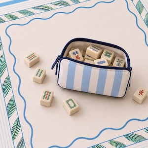 May include: A rectangular zippered pouch with light blue and white vertical stripes containing several off-white mahjong tiles with various symbols. Additional mahjong tiles are scattered on a white mat featuring a blue wavy border and green leaves.