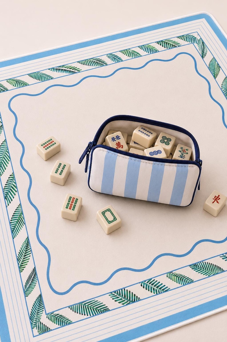 May include: A rectangular zippered pouch with light blue and white vertical stripes containing several off-white mahjong tiles with various symbols. Additional mahjong tiles are scattered on a white mat featuring a blue wavy border and green leaves.