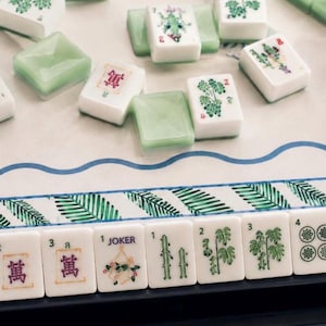 May include: A Mahjong set featuring white tiles with green and purple botanical and numerical designs, including a Joker tile. The tiles are arranged on a game board with a blue wavy border and a green palm leaf pattern, with some tiles in a black rack.