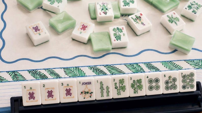 May include: A Mahjong set featuring white tiles with green and purple botanical and numerical designs, including a Joker tile. The tiles are arranged on a game board with a blue wavy border and a green palm leaf pattern, with some tiles in a black rack.