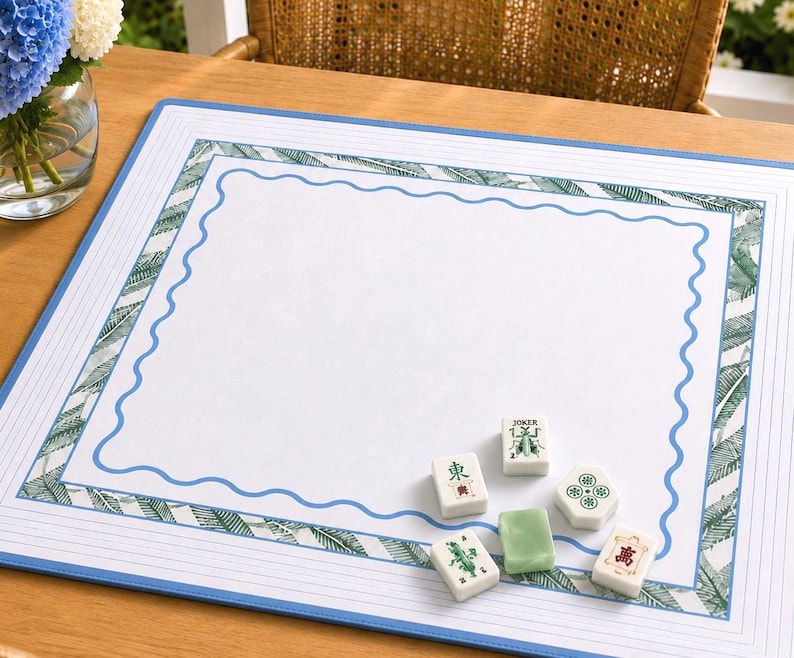 Coastal inspired mahjong mat on a sunlit outdoor table with botanical sage mahjong tiles and woven rattan chair backdrop