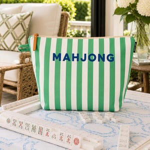 May include: A zipped pouch featuring vertical green and white stripes with the word MAHJONG printed in dark blue letters on the front. The pouch includes a small brown leather pull tab on the side zip and a black fabric brand tag.