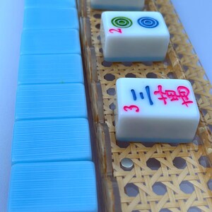 May include: A set of mahjong tiles with light blue backs and white fronts, featuring red, blue, and green engraved symbols and numbers. The tiles are arranged in a vertical row and on a clear rack with a woven tan cane pattern insert.