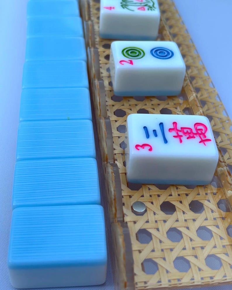 May include: A set of mahjong tiles with light blue backs and white fronts, featuring red, blue, and green engraved symbols and numbers. The tiles are arranged in a vertical row and on a clear rack with a woven tan cane pattern insert.