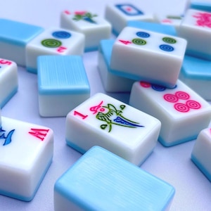May include: A collection of white Mahjong tiles with light blue backs, featuring various colorful engraved symbols, numbers, and Chinese characters in red, blue, green, and pink ink, scattered across a plain, light-colored surface.