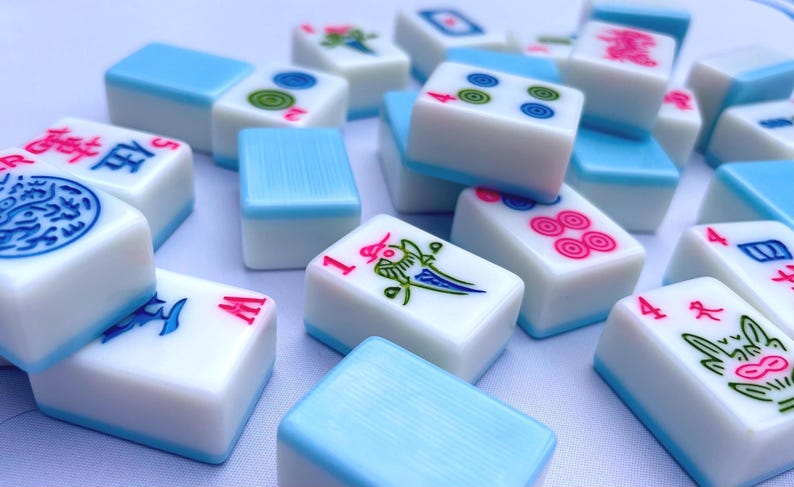 May include: A collection of white Mahjong tiles with light blue backs, featuring various colorful engraved symbols, numbers, and Chinese characters in red, blue, green, and pink ink, scattered across a plain, light-colored surface.