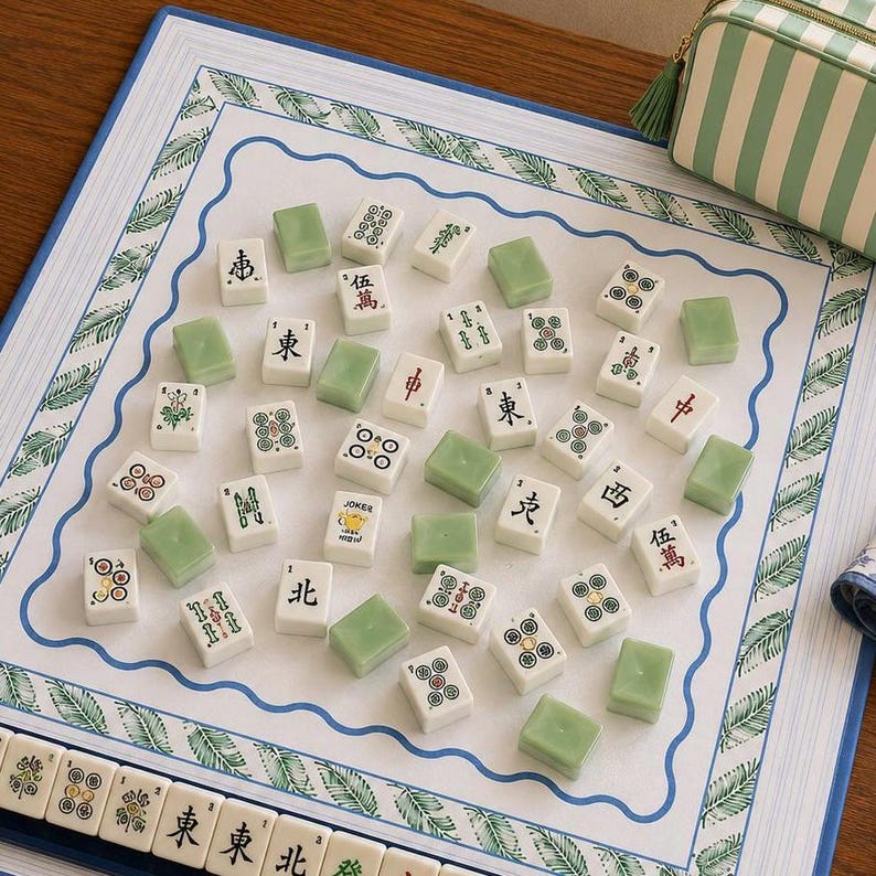 May include: A mahjong set featuring white tiles with black and green characters, symbols, and dots, alongside solid light green tiles. The tiles are arranged on a white play mat with a blue wavy border and green leaf patterns, with a striped pouch nearby.