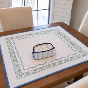 May include: A rectangular white fabric mat with a blue wavy border and a green leaf-patterned frame, featuring a small light blue and white striped zippered pouch containing several white rectangular tiles with blue and green markings.