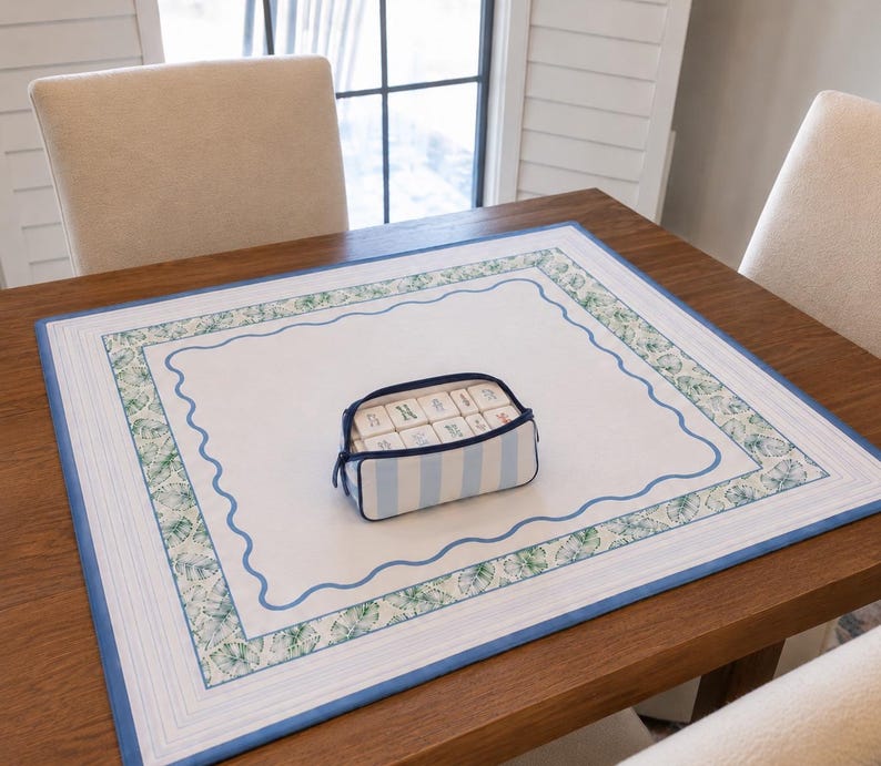 May include: A rectangular white fabric mat with a blue wavy border and a green leaf-patterned frame, featuring a small light blue and white striped zippered pouch containing several white rectangular tiles with blue and green markings.