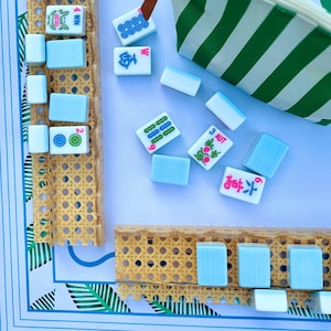 May include: A set of light blue and white Mahjong tiles with various colorful symbols and text including 4 WIN, 3 AUT, and 6, arranged on two tan woven cane racks. A green and white striped pouch sits nearby on a white surface with blue borders.