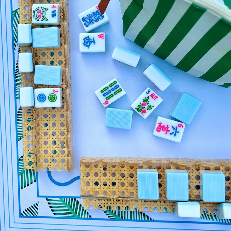May include: A set of light blue and white Mahjong tiles with various colorful symbols and text including 4 WIN, 3 AUT, and 6, arranged on two tan woven cane racks. A green and white striped pouch sits nearby on a white surface with blue borders.