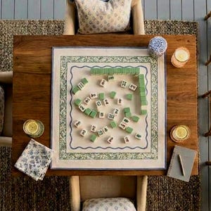 May include: A square mahjong game mat with a green and blue floral border, featuring scattered white tiles with black characters and green-backed tiles arranged on a wooden table, accompanied by two upholstered chairs with patterned pillows.