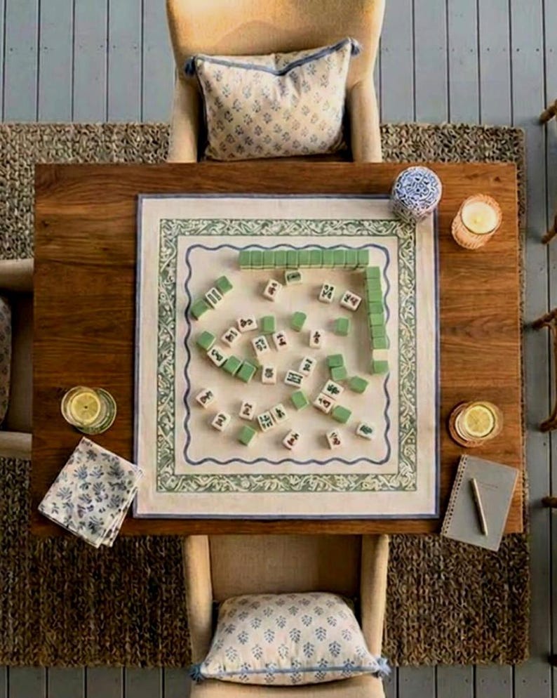 May include: A square mahjong game mat with a green and blue floral border, featuring scattered white tiles with black characters and green-backed tiles arranged on a wooden table, accompanied by two upholstered chairs with patterned pillows.