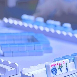 May include: A Mahjong set featuring light blue tiles arranged in racks and a central square formation. The tiles display various symbols, numbers, and the word JOKER in red and blue ink. The game components are uniform in color and design.