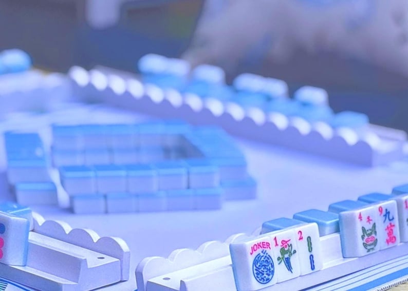 May include: A Mahjong set featuring light blue tiles arranged in racks and a central square formation. The tiles display various symbols, numbers, and the word JOKER in red and blue ink. The game components are uniform in color and design.