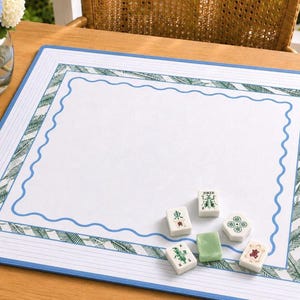 May include: A rectangular mahjong mat featuring a white center with a blue wavy border, surrounded by a green palm leaf pattern and a solid blue outer edge. Six mahjong tiles are placed on the mat, including one blank green tile.