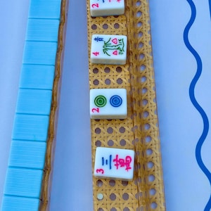 May include: A set of mahjong tiles and two long, rectangular racks with a woven cane texture. One rack holds a row of light blue tiles, while the other displays four individual tiles featuring red, green, and blue symbols and Chinese characters.