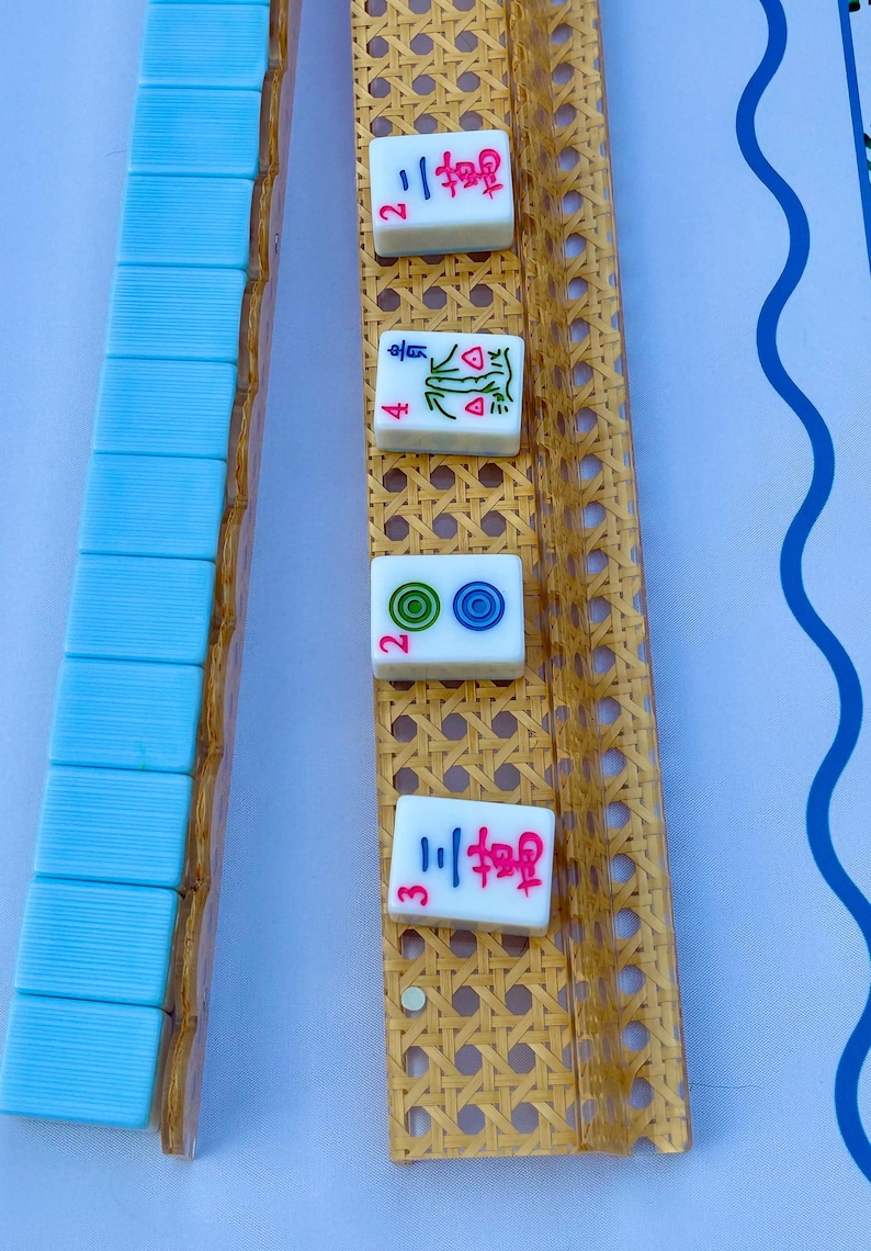 May include: A set of mahjong tiles and two long, rectangular racks with a woven cane texture. One rack holds a row of light blue tiles, while the other displays four individual tiles featuring red, green, and blue symbols and Chinese characters.