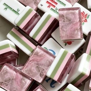 May include: A collection of rectangular mahjong tiles featuring a mix of designs. Some tiles are white with green bamboo or red floral motifs, while others display horizontal stripes of white, green, and pink, or translucent pink crystalline textures.