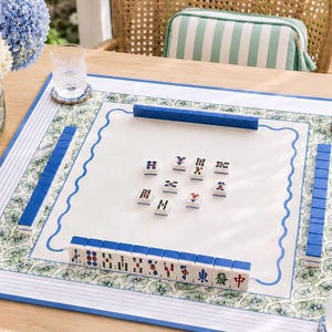 May include: A square mahjong mat featuring a white center with a blue wavy border, surrounded by a green and white botanical print and blue striped edges. The mat displays rows of blue-backed mahjong tiles and several tiles placed in the center.