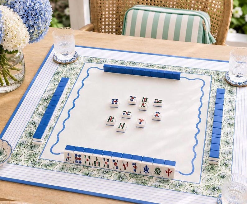 May include: A square mahjong mat featuring a white center with a blue wavy border, surrounded by a green and white botanical print and blue striped edges. The mat displays rows of blue-backed mahjong tiles and several tiles placed in the center.