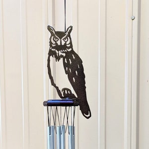 May include: A metal wind chime featuring a black silhouette of an owl perched on a brown wooden platform. The chime has silver tubes and a brown wooden bead. The owl is facing to the right and has a detailed design.