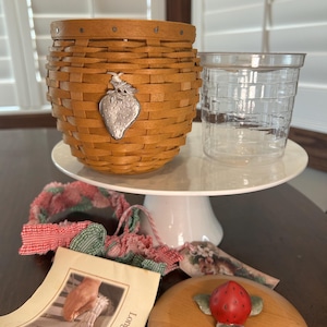 May include: A woven basket with a silver strawberry charm, a clear plastic cup, and a wooden lid with a red strawberry on top. The basket and cup sit on a white cake stand. A small booklet and fabric accents are also present.