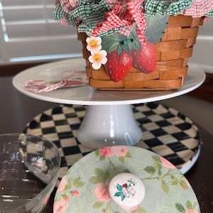 May include: A decorative picnic basket with a red and green gingham fabric lining and strawberry accents. The basket sits on a white cake stand, with a floral-patterned plate and a clear glass bowl in the foreground. The basket is made of woven wood.