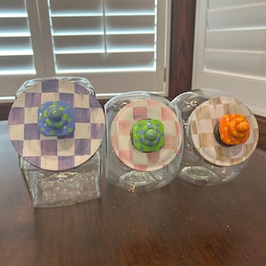 May include: Three clear glass jars with square bases and round lids. Each lid features a hand-painted checkered pattern in purple, pink, or tan. Centered on each lid is a small, colorful, sculpted knob with polka dots in blue, green, or orange.