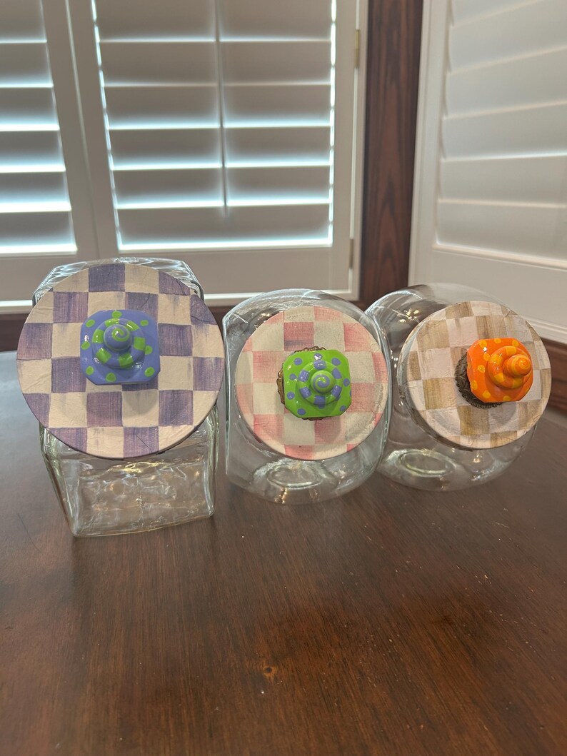 May include: Three clear glass jars with square bases and round lids. Each lid features a hand-painted checkered pattern in purple, pink, or tan. Centered on each lid is a small, colorful, sculpted knob with polka dots in blue, green, or orange.