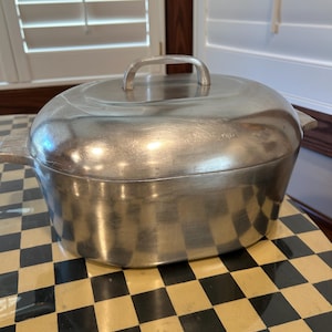 May include: An oval-shaped silver-toned metal roasting pan with a matching lid. The cookware features a smooth, brushed finish, a central handle on the lid, and integrated side handles on the main body of the pot for easy lifting.
