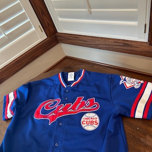 May include: A royal blue Chicago Cubs baseball jersey featuring a red and white script logo across the chest, a circular team patch on the lower front, and a commemorative patch on the sleeve. The shirt has a button-down front and striped trim.