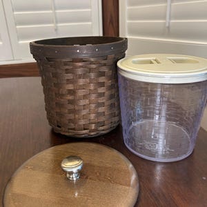 May include: A woven brown basket, a clear plastic container with a white lid, and a wooden lid with a silver knob are displayed on a dark wooden surface. The basket has a dark rim and a woven pattern. The container is cylindrical.