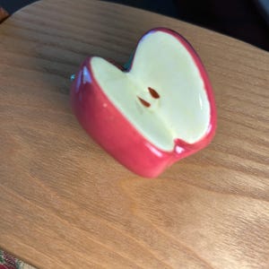 May include: A ceramic apple-shaped decorative item. The apple is cut in half, revealing a creamy white interior with two brown seeds. The exterior is a glossy red. The item rests on a wooden surface.