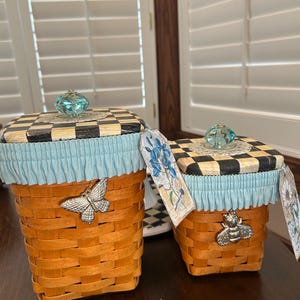May include: Two decorative woven baskets with black and white checkered lids and light blue ruffled trim. Each basket has a silver-colored metal charm, one a butterfly and the other a bee. The lids are topped with a clear, faceted knob.