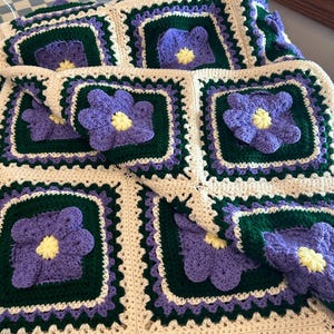 May include: A handmade crochet blanket featuring a repeating pattern of square motifs. Each square consists of a central purple flower with a yellow center, surrounded by a dark green border and an outer frame of cream-colored yarn.